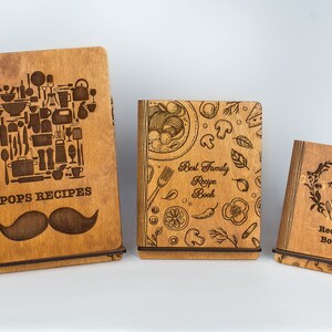 May include: Three wooden recipe books with engraved designs. The first book has a chef's hat made of kitchen utensils and the text "Pops Recipes" with a mustache. The second book has a design of food and the text "Best Family Recipe Book". The third book has a wreath with the text "Maria's Recipe Book".