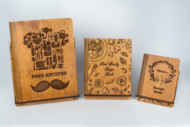 May include: Three wooden recipe books with engraved designs. The first book has a chef's hat made of kitchen utensils and the text "Pops Recipes" with a mustache. The second book has a design of food and the text "Best Family Recipe Book". The third book has a wreath with the text "Maria's Recipe Book".