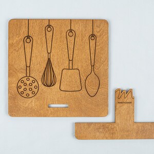 May include: Wooden kitchen utensil holder with a cutout design of five hanging utensils. The holder has a small, rectangular cutout on the bottom and the text "MARKITSHOP" is engraved on the side.