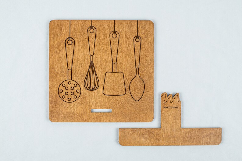 May include: Wooden kitchen utensil holder with a cutout design of five hanging utensils. The holder has a small, rectangular cutout on the bottom and the text "MARKITSHOP" is engraved on the side.