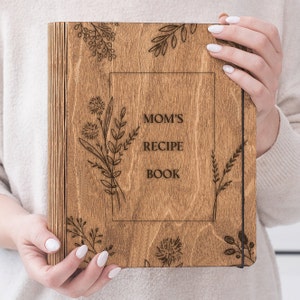 Peut inclure: Un livre de recettes en bois avec une couverture en bois brun et un design grav&eacute; au laser. La couverture pr&eacute;sente le texte "Mom's Recipe Book" et un motif floral.