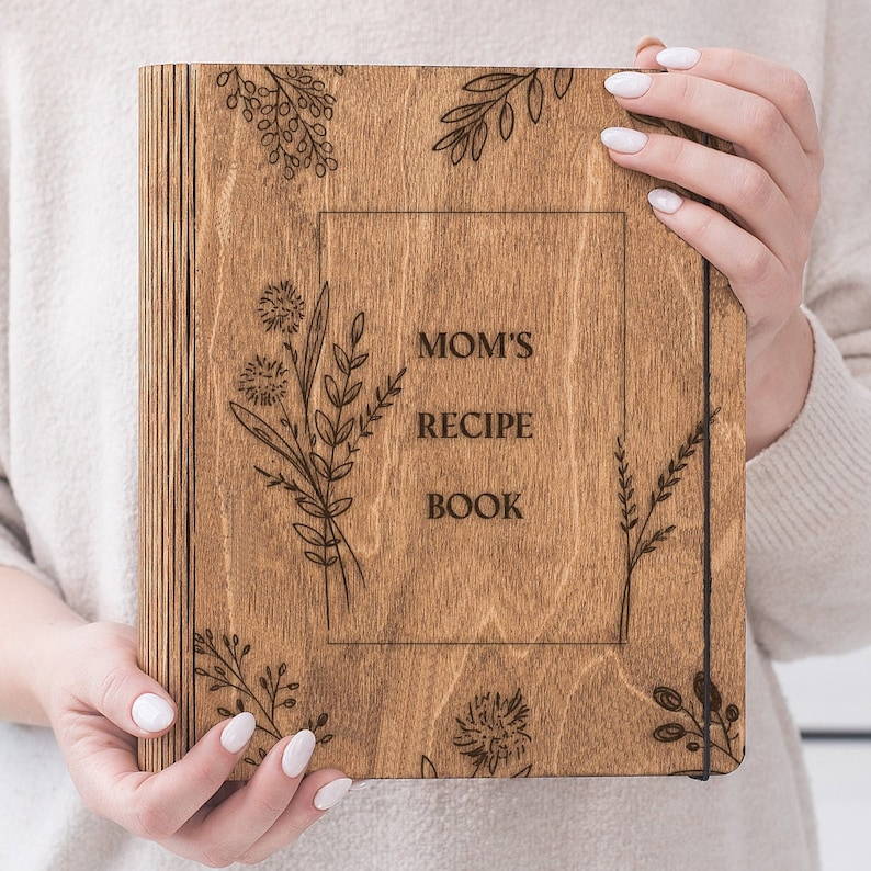 Peut inclure: Un livre de recettes en bois avec une couverture en bois brun et un design grav&eacute; au laser. La couverture pr&eacute;sente le texte "Mom's Recipe Book" et un motif floral.