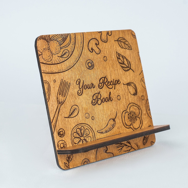May include: A wooden recipe book stand with a brown wood finish. The stand has a cutout design with a fork, chili peppers, lemons, and other food-related illustrations. The text "Your Recipe Book" is engraved on the stand.