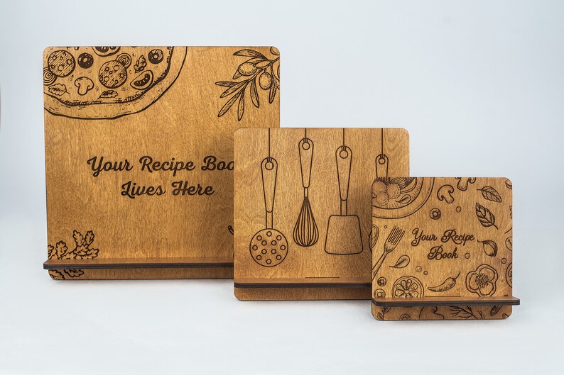 May include: Three wooden recipe book stands of different sizes. The largest stand has the text "Your Recipe Book Lives Here" and a pizza graphic. The middle stand has a graphic of kitchen utensils. The smallest stand has a graphic of various food items and the text "Your Recipe Book".