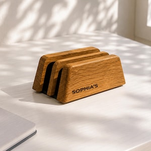 May include: A wooden vertical laptop stand with a natural grain finish and two slots for holding devices. The front face of the stand features the engraved text SOPHIA'S in a dark, sans-serif font. The stand has a trapezoidal profile.