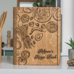 Peut inclure: Un livre de recettes en bois avec une couverture en bois brun et un motif grav&eacute; au laser de l&eacute;gumes et d'herbes. Le texte "Rebecca's Recipe Book" est grav&eacute; sur la couverture.