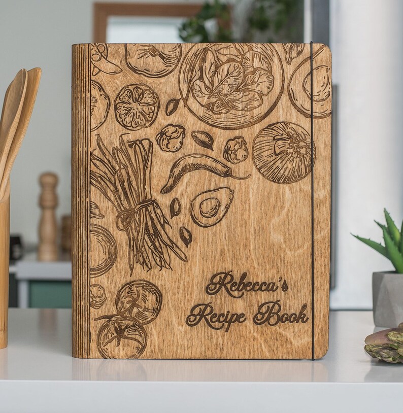 Peut inclure: Un livre de recettes en bois avec une couverture en bois brun et un motif grav&eacute; au laser de l&eacute;gumes et d'herbes. Le texte "Rebecca's Recipe Book" est grav&eacute; sur la couverture.