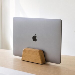 May include: A silver laptop with a black apple logo on the back is held in an upright, vertical position by a solid, light-coloured wooden stand. The stand has a trapezoidal shape with rounded edges and a smooth, natural wood grain finish.