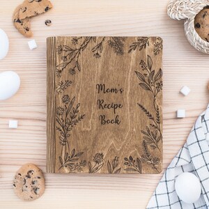 Puede incluir: Un libro de recetas de madera con un dise&ntilde;o floral grabado en la portada. El libro se titula "Mom's Recipe Book". El libro est&aacute; rodeado de galletas, terrones de az&uacute;car y huevos sobre una superficie de madera.