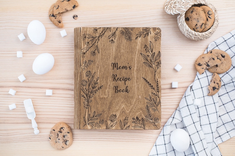 Puede incluir: Un libro de recetas de madera con un dise&ntilde;o floral grabado en la portada. El libro se titula "Mom's Recipe Book". El libro est&aacute; rodeado de galletas, terrones de az&uacute;car y huevos sobre una superficie de madera.