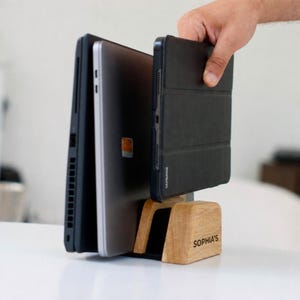 May include: A light-colored wooden vertical laptop and tablet stand featuring the name SOPHIA'S engraved on the front. The stand holds a black laptop, a silver laptop, and a black tablet in separate slots, providing a stable base for devices.