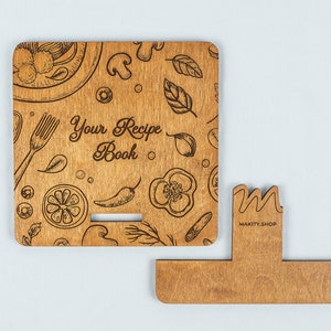 May include: Wooden recipe book stand with a brown wood finish. The stand has a cutout for a book and a decorative front panel with a food-themed design and the text "Your Recipe Book".