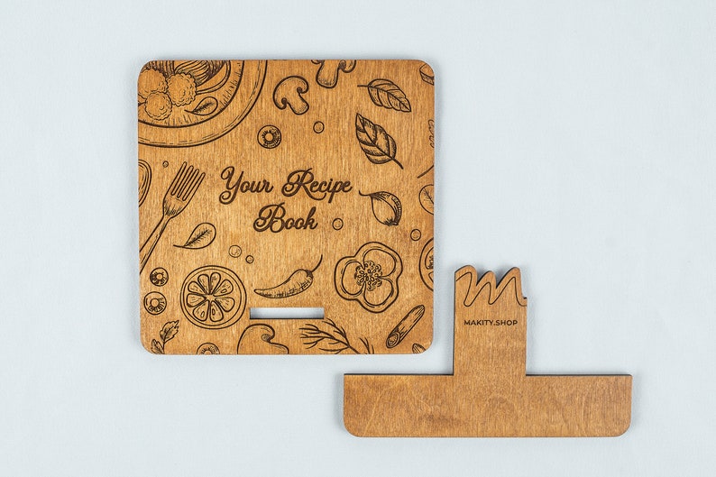 May include: Wooden recipe book stand with a brown wood finish. The stand has a cutout for a book and a decorative front panel with a food-themed design and the text "Your Recipe Book".