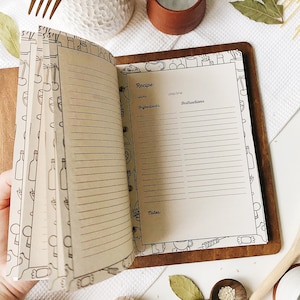 Puede incluir: Libro de recetas abierto con p&aacute;ginas en blanco para escribir recetas. Las p&aacute;ginas est&aacute;n rayadas y tienen un borde decorativo de ilustraciones con tem&aacute;tica de cocina. El libro est&aacute; encuadernado en cuero marr&oacute;n y tiene una cubierta de madera.
