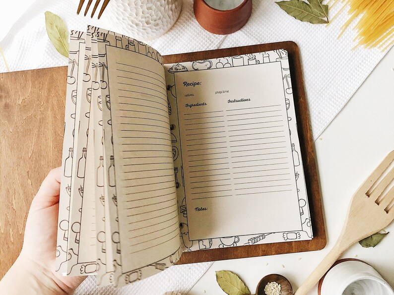 Puede incluir: Libro de recetas abierto con p&aacute;ginas en blanco para escribir recetas. Las p&aacute;ginas est&aacute;n rayadas y tienen un borde decorativo de ilustraciones con tem&aacute;tica de cocina. El libro est&aacute; encuadernado en cuero marr&oacute;n y tiene una cubierta de madera.