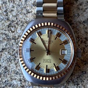 Puede incluir: Un reloj de pulsera de dos tonos, dorado y plateado, con una esfera redonda y un bisel dorado. La esfera del reloj es blanca con marcadores de hora romanos negros y agujas negras. El reloj tiene una ventana de fecha en la posición de las 3 en punto y el texto "Savar 25 Rubis" y "Automatic Incabloc 10 Atmos" impreso en la esfera. El reloj está en una banda de metal de dos tonos, dorado y plateado.