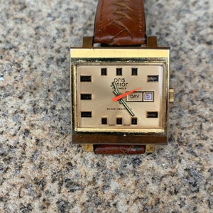 Puede incluir: Un reloj de pulsera cuadrado dorado con una correa de cuero marrón. La esfera del reloj es dorada con marcas negras y el texto "Oris Junior 17 Jewels" y "Shock-Resist". El día de la semana se muestra como "DAY" y la fecha es "5".