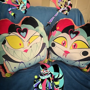May include: Two colorful pillow covers featuring a cartoon character with a large grin and a heart on its forehead. The character is wearing a jester hat and has a pink and blue color scheme. The pillows are on a blue couch with a third pillow cover featuring the same character in a different pose.