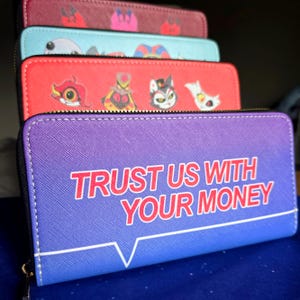 May include: A stack of zippered wallets in various colors. The front wallet is purple with the text "TRUST US WITH YOUR MONEY" in red. Other wallets are red, teal, and maroon, each with cartoon character designs.