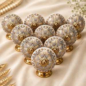 May include: A collection of ten round ceramic cabinet knobs arranged in a triangular formation. Each knob features a grey and white patterned surface with gold-toned accents and a polished gold-toned metal base at the centre and bottom.