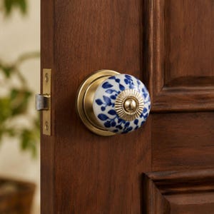 Op de afbeelding: Een ronde keramische deurknop met een witte basis en een blauw bladpatroon. De knop heeft een centrale metalen dop in messingkleur met een geribbelde textuur en is gemonteerd op een ronde metalen plaat op een donkerbruine houten deur.