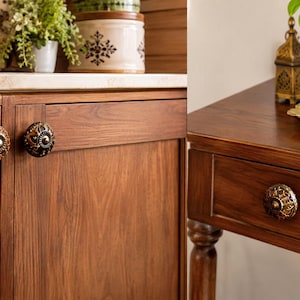May include: Round cabinet knobs with intricate metallic gold and dark brown floral patterns. These circular hardware pieces are mounted on wooden cabinet doors and a wooden drawer, featuring a central raised button and detailed embossed edges.