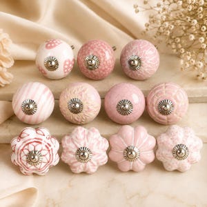 May include: A collection of twelve ceramic cabinet knobs in various shades of pink and white. The knobs feature diverse patterns including hearts, stripes, floral motifs, and crackled textures, each finished with a central silver metal screw.