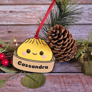 May include: A yellow dumpling-shaped ornament with a smiling face and pink cheeks, sitting in a tan steamer basket base labelled with the name Cassandra in black text. The ornament hangs from a thin red ribbon against a festive holiday backdrop.