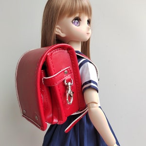 May include: A red school backpack with a silver clasp and white trim. The backpack is worn by a doll with long brown hair and purple eyes, wearing a white and blue sailor uniform and a navy blue skirt.