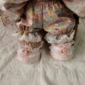 Puede incluir: Una mu&ntilde;eca con un vestido de estampado floral y bombachos a juego con ribete de encaje. La mu&ntilde;eca lleva zapatos de rayas rosa y blancas con un adorno de flor rosa.