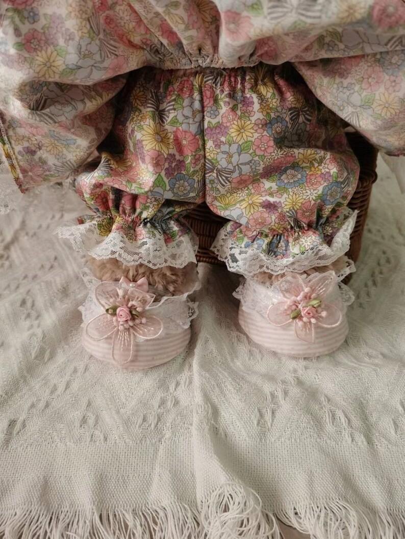 Puede incluir: Una mu&ntilde;eca con un vestido de estampado floral y bombachos a juego con ribete de encaje. La mu&ntilde;eca lleva zapatos de rayas rosa y blancas con un adorno de flor rosa.