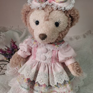 Puede incluir: Un oso de peluche marr&oacute;n que lleva un vestido a rayas rosa y blanco con ribete de encaje y una diadema floral. El vestido tiene un lazo rosa y botones.
