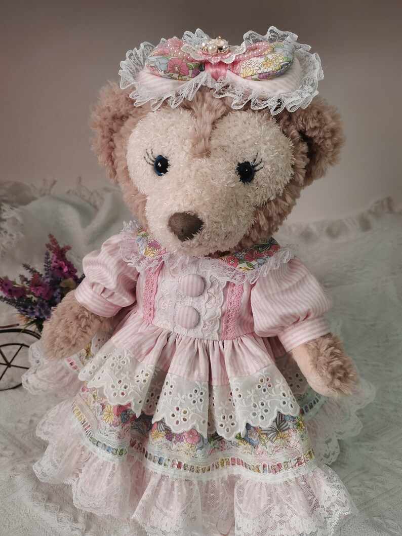 Puede incluir: Un oso de peluche marr&oacute;n que lleva un vestido a rayas rosa y blanco con ribete de encaje y una diadema floral. El vestido tiene un lazo rosa y botones.