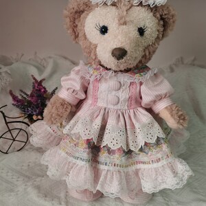 Puede incluir: Un oso de peluche que lleva un vestido rosa y blanco con ribete de encaje y estampado floral. El oso lleva un sombrero rosa y blanco con un lazo y una perla.