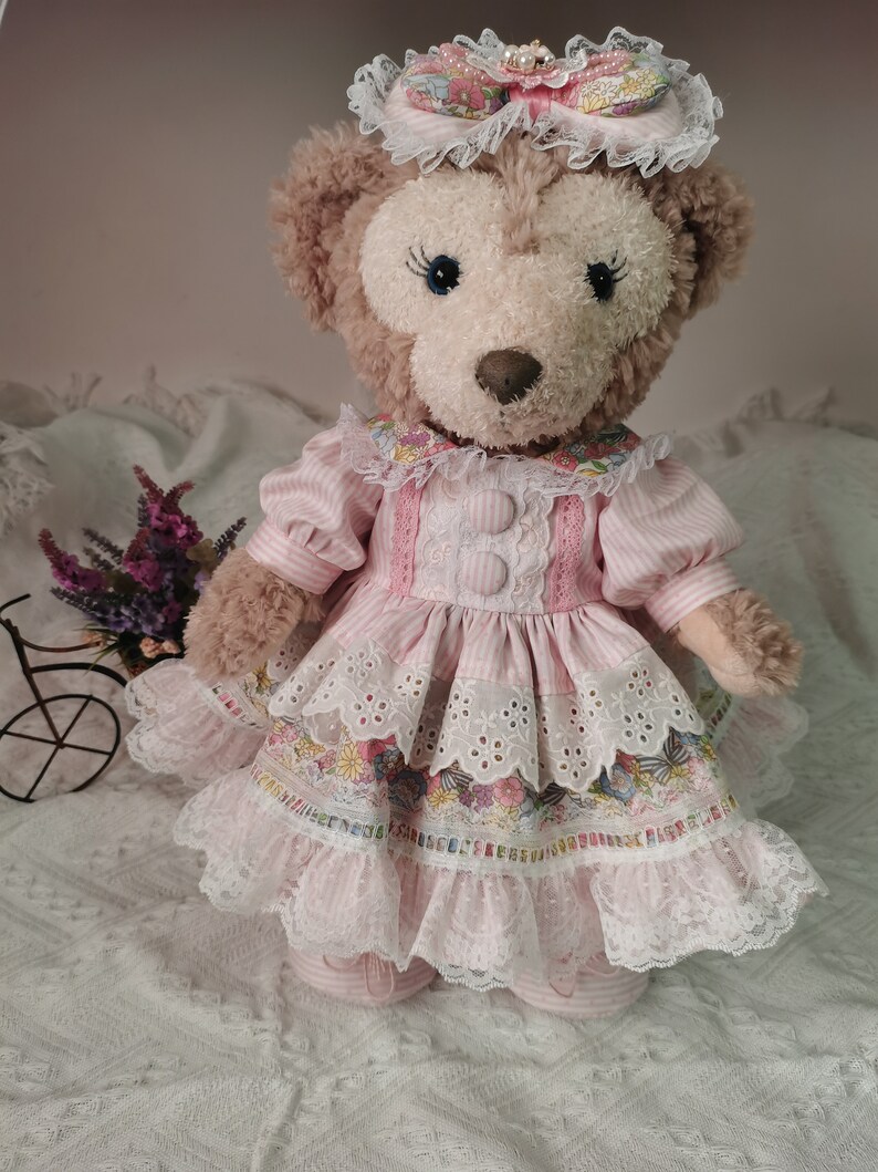 Puede incluir: Un oso de peluche que lleva un vestido rosa y blanco con ribete de encaje y estampado floral. El oso lleva un sombrero rosa y blanco con un lazo y una perla.