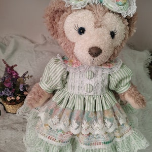 Puede incluir: Un oso de peluche marr&oacute;n que lleva un vestido a rayas verdes y blancas con ribetes de encaje y una diadema floral. El vestido tiene una falda de capas con un estampado floral y detalles de encaje.