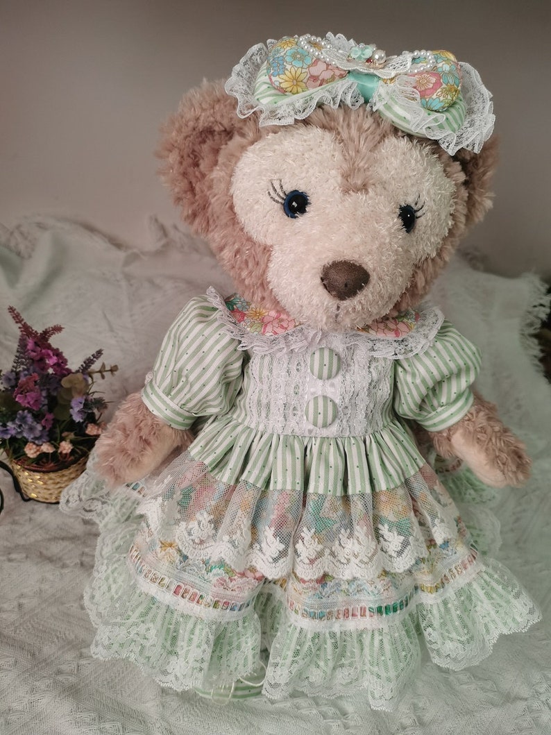 Puede incluir: Un oso de peluche marr&oacute;n que lleva un vestido a rayas verdes y blancas con ribetes de encaje y una diadema floral. El vestido tiene una falda de capas con un estampado floral y detalles de encaje.