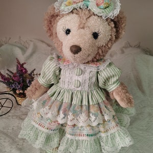 Puede incluir: Un oso de peluche marr&oacute;n que lleva un vestido a rayas verdes y blancas con ribetes de encaje y una diadema floral.