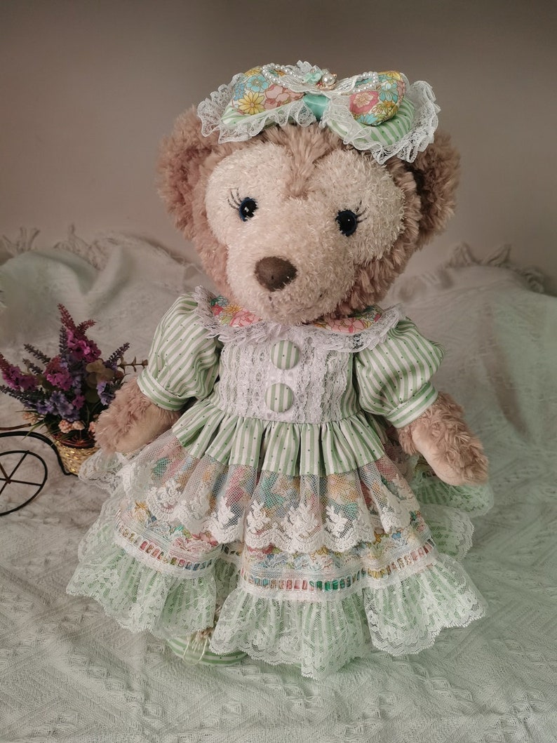 Puede incluir: Un oso de peluche marr&oacute;n que lleva un vestido a rayas verdes y blancas con ribetes de encaje y una diadema floral.