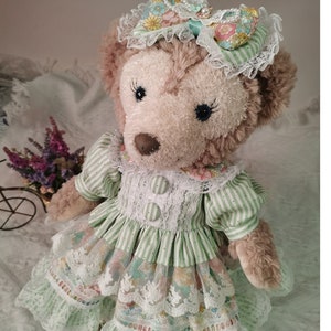 Puede incluir: Un oso de peluche marr&oacute;n que lleva un vestido a rayas verdes y blancas con estampado floral y ribete de encaje. El oso lleva un sombrero de encaje blanco con un lazo floral y detalles de perlas.