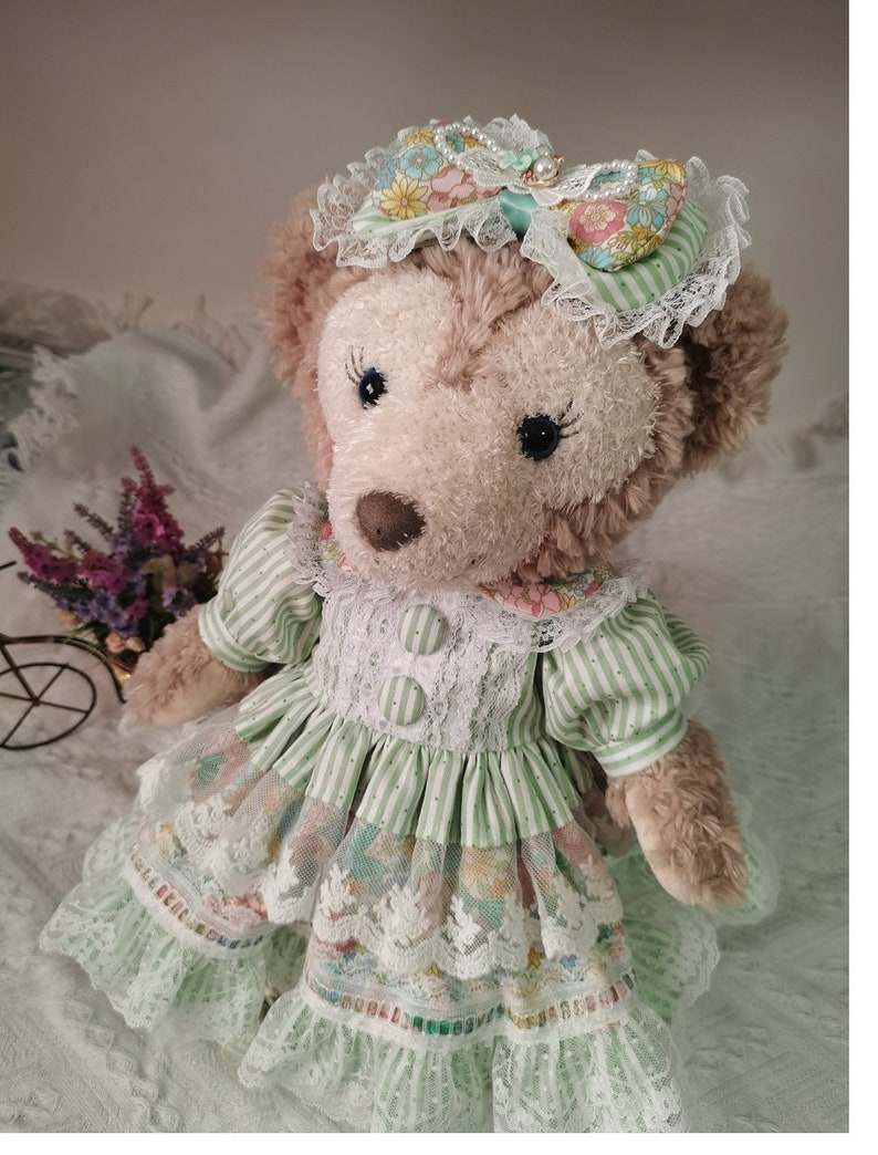 Puede incluir: Un oso de peluche marr&oacute;n que lleva un vestido a rayas verdes y blancas con estampado floral y ribete de encaje. El oso lleva un sombrero de encaje blanco con un lazo floral y detalles de perlas.