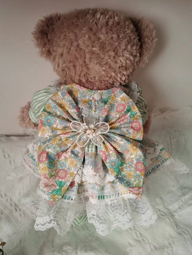 Puede incluir: Un oso de peluche marr&oacute;n que lleva un vestido a rayas verdes y blancas con estampado floral y un gran lazo blanco. El vestido tiene un borde de encaje y un bot&oacute;n blanco en la espalda.