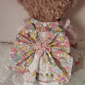 Puede incluir: Un vestido de estampado floral rosa y blanco con un gran lazo rosa y ribete de encaje blanco. El vestido lo lleva un oso de peluche marr&oacute;n.