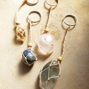 Könnte beinhalten: Fünf Edelstein-Schlüsselanhänger mit silbernen Ringen und natürlichen Schnurkordeln. Die Edelsteine sind in verschiedenen Farben erhältlich, darunter Gelb, Blau, Rosa und Grün. Jeder Stein ist in einem Makramee-Netz mit einem Holzperlenakzent eingeschlossen.