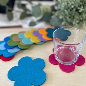Könnte beinhalten: Ein Set aus bunten Filz-Untersetzern in Blumenform. Die Untersetzer sind in einem Regenbogenmuster angeordnet, mit einem Glas auf einem rosafarbenen Untersetzer.