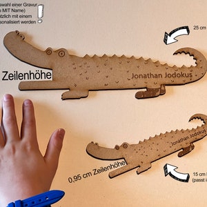 Może przedstawiać: Dwie drewniane zakładki w kształcie krokodyla. Większa ma 25 cm długości, a mniejsza 15 cm. Obie mają wygrawerowane imię "Jonathan Jodokus". Większa zakładka ma 1,5 cm wysokości, a mniejsza 0,95 cm.