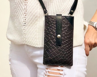 May include: A black leather phone case with a crocodile pattern and a silver button. The case is attached to a black strap and worn across the body.