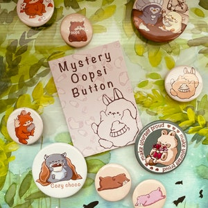 May include: A collection of 10 round button pins featuring various cartoon animals. The buttons are white with colorful illustrations. The buttons are arranged on a green and brown background with watercolor-like designs. The text "Mystery Oopsi Button" is printed on a pink card.