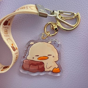 Może przedstawiać: Brelok z przezroczystego akrylu z kreskówkową ilustracją śpiącego żółtego kurczaka na brązowej poduszce. Brelok ma złoty metalowy zamek i biało-brązowy pasek z nadrukiem "Puddings".