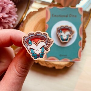 Op de afbeelding: Een houten pin met een cartoon gezicht dat huilt. De pin is geschilderd in blauw, rood en wit. De pin is gelabeld "Emotionele Pins".